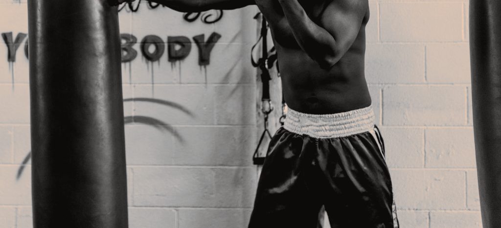 A boxer's bare torso between two heavy bags, hands raised in a guard, in front of a painted gym wall